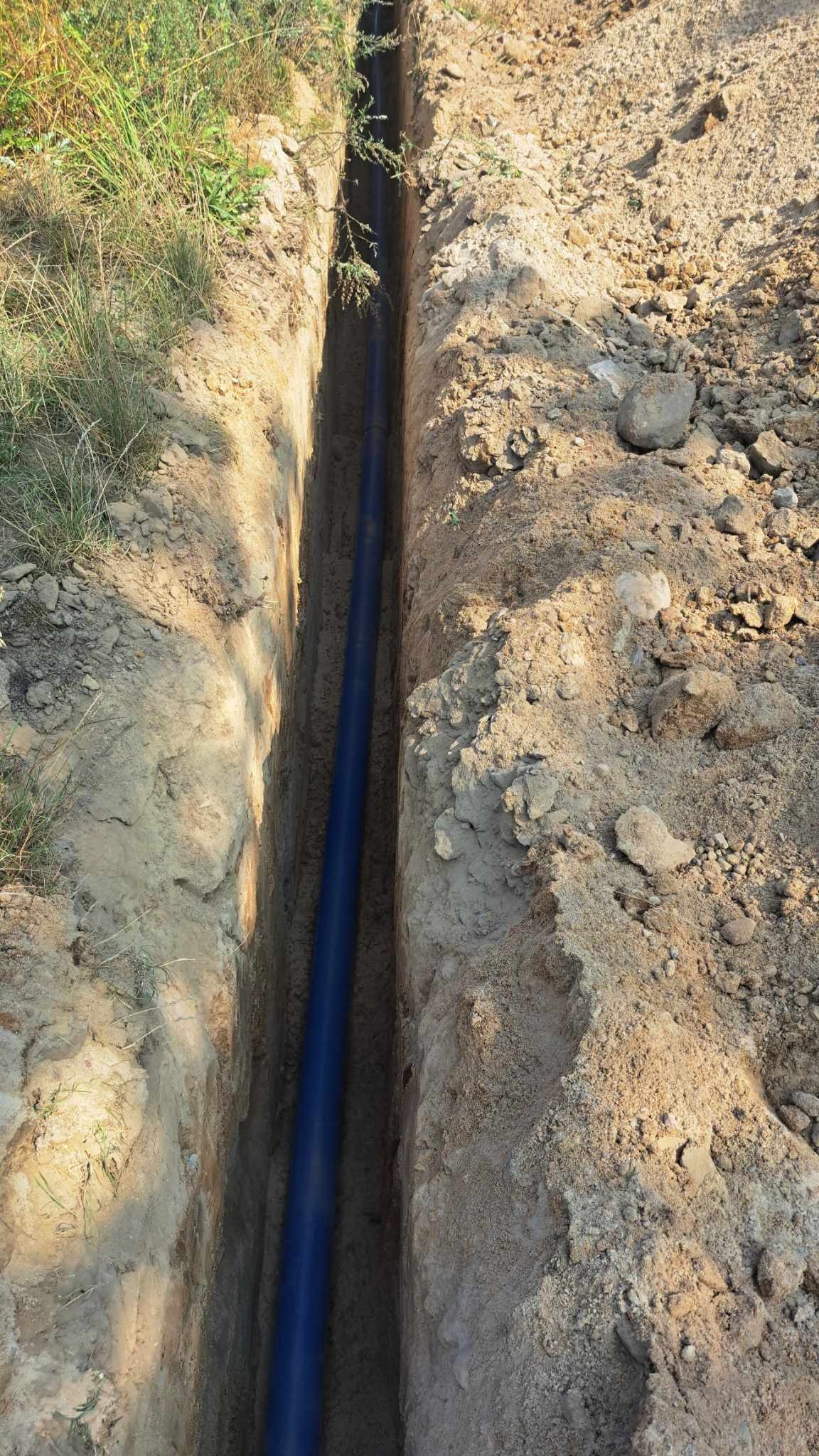 Długi, niebieski rurociąg ułożony w wąskim wykopie ziemnym, otoczony krawędziami z piasku i ziemi, z zieloną trawą rosnącą wzdłuż jednego brzegu wykopu.