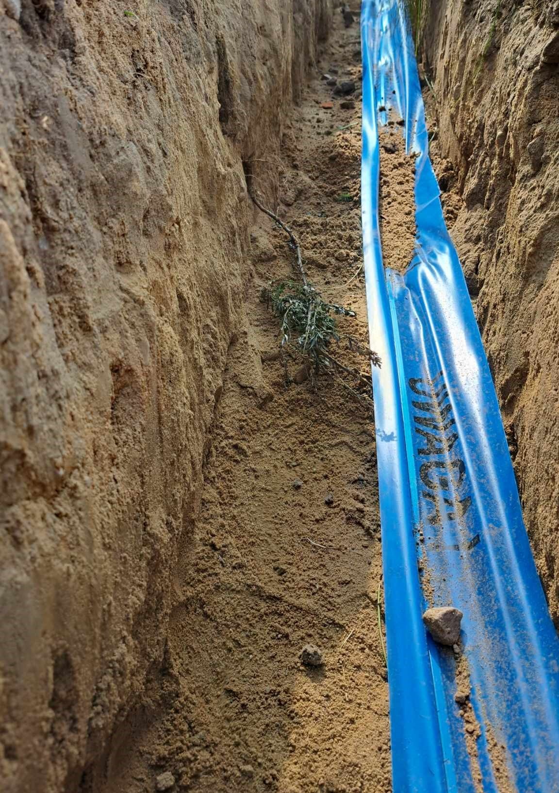 Wykop z piaskowymi ścianami, w którym ułożono niebieską rurę osłonową z widocznym napisem, częściowo zasypaną piaskiem.