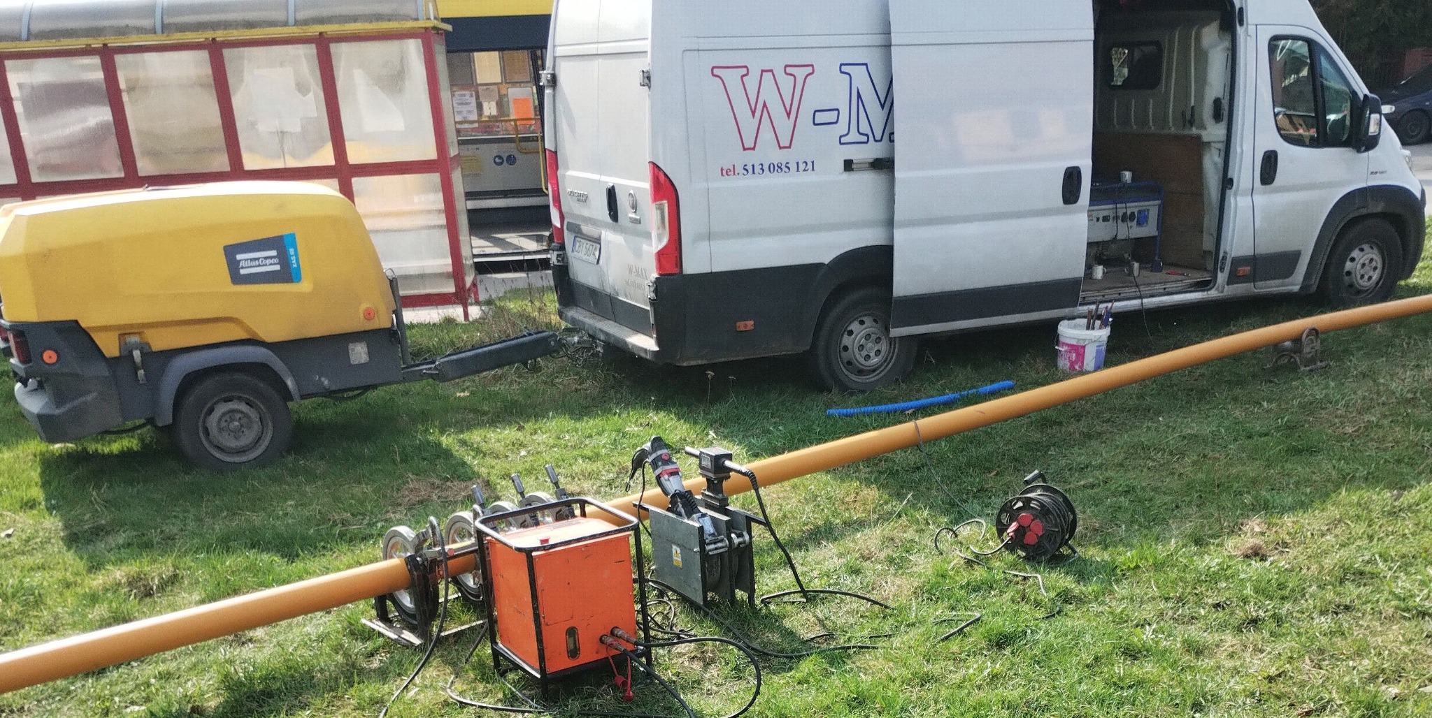 Zgrzewanie rury z tworzywa sztucznego na trawie, widoczny agregat prądotwórczy na przyczepce, samochód dostawczy z otwartymi drzwiami oraz zgrzewarka. Ujęcie z bliska, w tle przystanek autobusowy.