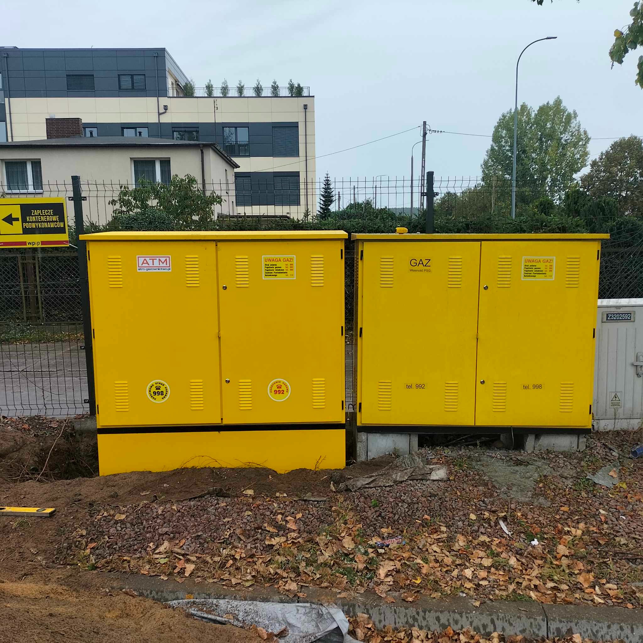 Dwa żółte, metalowe regulatory gazu na zewnątrz, z naklejkami ostrzegawczymi i numerami telefonów alarmowych, ustawione obok siebie na żwirowym podłożu, z budynkami mieszkalnymi w tle.