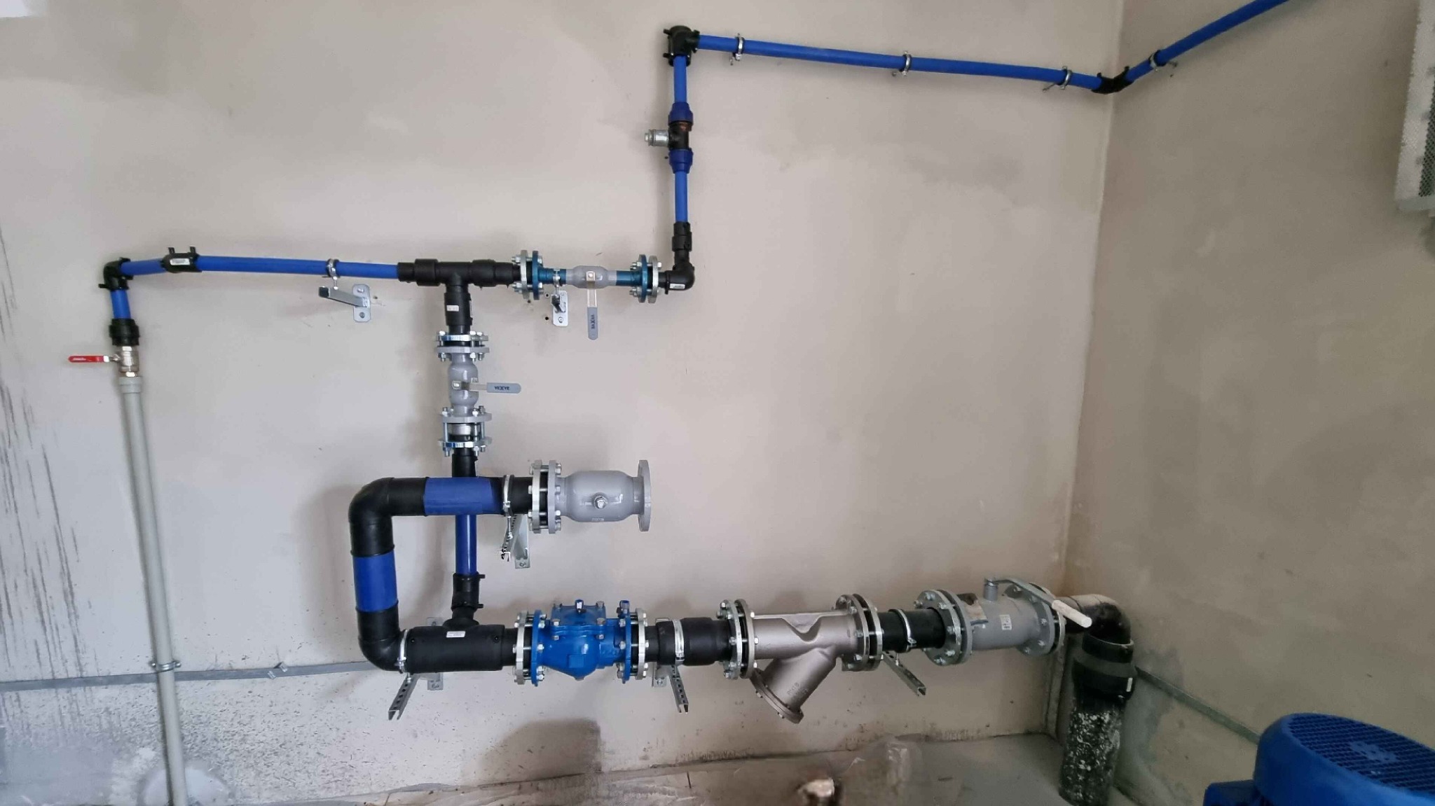Złożona instalacja wodna z rur niebieskich i czarnych, z licznymi zaworami i filtrami, zamontowana na surowej, szarej ścianie w pomieszczeniu technicznym.