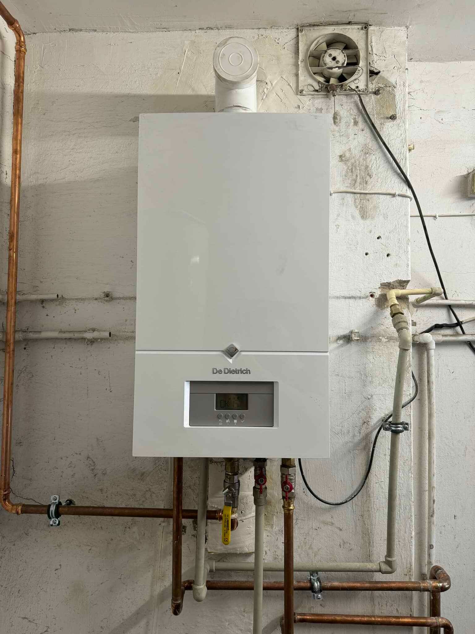 Biały, wiszący piec gazowy De Dietrich na tle surowej, pomalowanej ściany z widocznymi rurami miedzianymi i plastikowymi, wentylator wyciągowy w górnym rogu, żółty zawór gazowy.