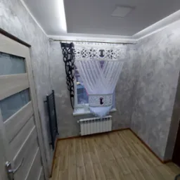 Pokój po remoncie: szpachlowane ściany w odcieniach szarości, drewnopodobna podłoga, biały grzejnik pod oknem z dekoracyjnymi firanami w czarno-białe wzory, drzwi wewnętrzne z przeszkleniami...