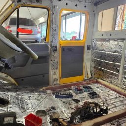 Versitale Kacper Pirch - Wnętrze demontowanego pojazdu dostawczego z żółtymi elementami karoserii, wygłuszone matami bitumicznymi, widoczne narzędzia i akcesoria warsztatowe.