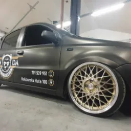 Matowy, czarny samochód z naklejonym logo 'PANDA AUTO SERWIS' i złotymi felgami typu mesh, zaparkowany w warsztacie.