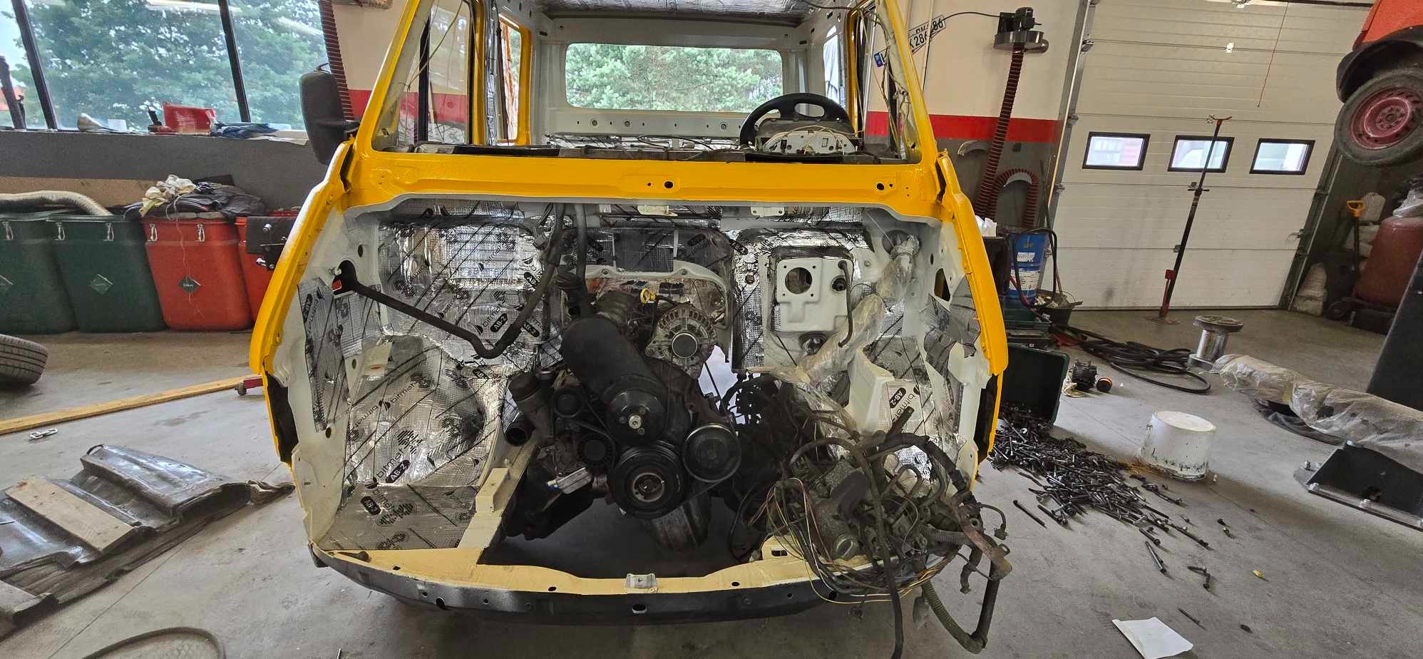 Żółty samochód dostawczy w trakcie renowacji, z widocznym silnikiem, wygłuszeniem ścianek bocznych, wnętrzem kabiny kierowcy i elementami karoserii w warsztacie.