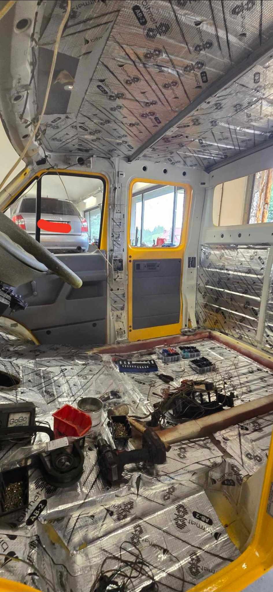 Wnętrze demontowanego pojazdu dostawczego z żółtymi elementami karoserii, wygłuszone matami bitumicznymi, widoczne narzędzia i akcesoria warsztatowe.