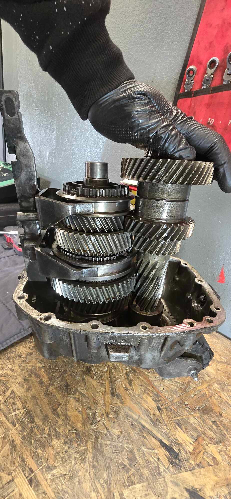 Otwarta skrzynia biegów samochodowych z widocznymi przekładniami, trzymana przez dłoń w czarnej rękawicy roboczej, na tle ściany warsztatu z narzędziami.