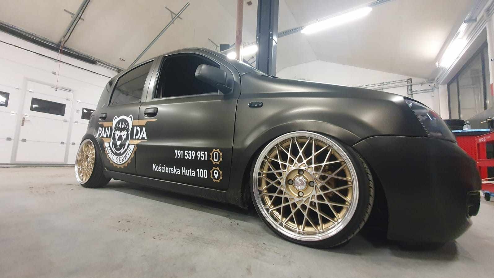 Matowy, czarny samochód z naklejonym logo 'PANDA AUTO SERWIS' i złotymi felgami typu mesh, zaparkowany w warsztacie.