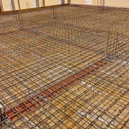 MBS BANKING MAGDALENA BARTKOWIAK-STEMPAK - Siatka zbrojeniowa ułożona na żółtym szalunku, przygotowana do zalania betonem na budowie, widoczne wystające pręty i fragment budynku z żółtą elewacją w tle.