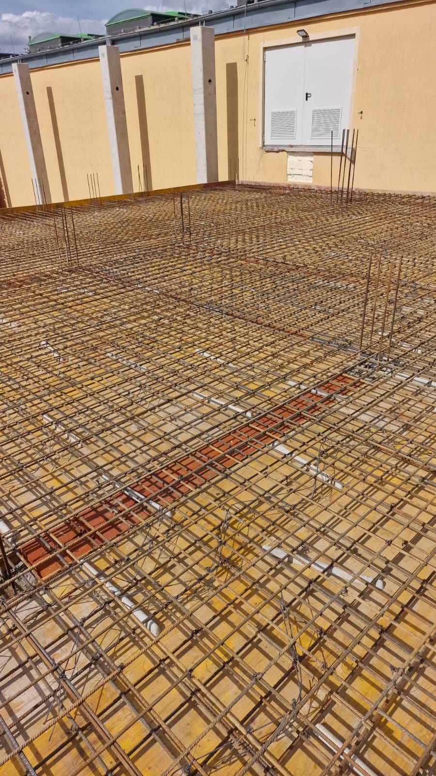 Siatka zbrojeniowa ułożona na żółtym szalunku, przygotowana do zalania betonem na budowie, widoczne wystające pręty i fragment budynku z żółtą elewacją w tle.