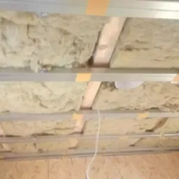 Montaż izolacji z wełny mineralnej w metalowym stelażu sufitu podwieszanego, widoczne drewniane elementy konstrukcyjne i taśma mocująca.