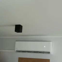 Biały klimatyzator ścienny z wyświetlaczem temperatury, zamontowany w pomieszczeniu w Tomaszowie Mazowieckim, widoczne fragmenty mebli i oświetlenie sufitowe.