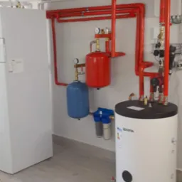 Nowoczesna kotłownia z białym zasobnikiem ciepłej wody, czerwonymi rurami w izolacji, białym piecem i niebieskim naczyniem przeponowym, uzupełniona filtrami wody.