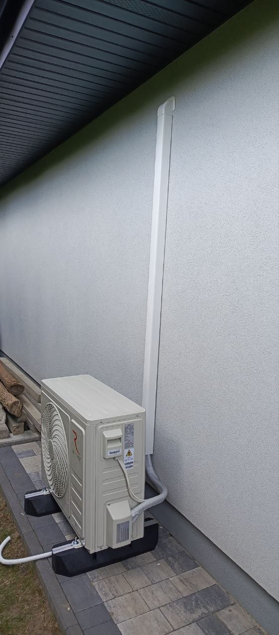 Zewnętrzna jednostka klimatyzacji zamontowana na czarnych podstawach antywibracyjnych, z widoczną białą listwą maskującą rury na elewacji budynku.