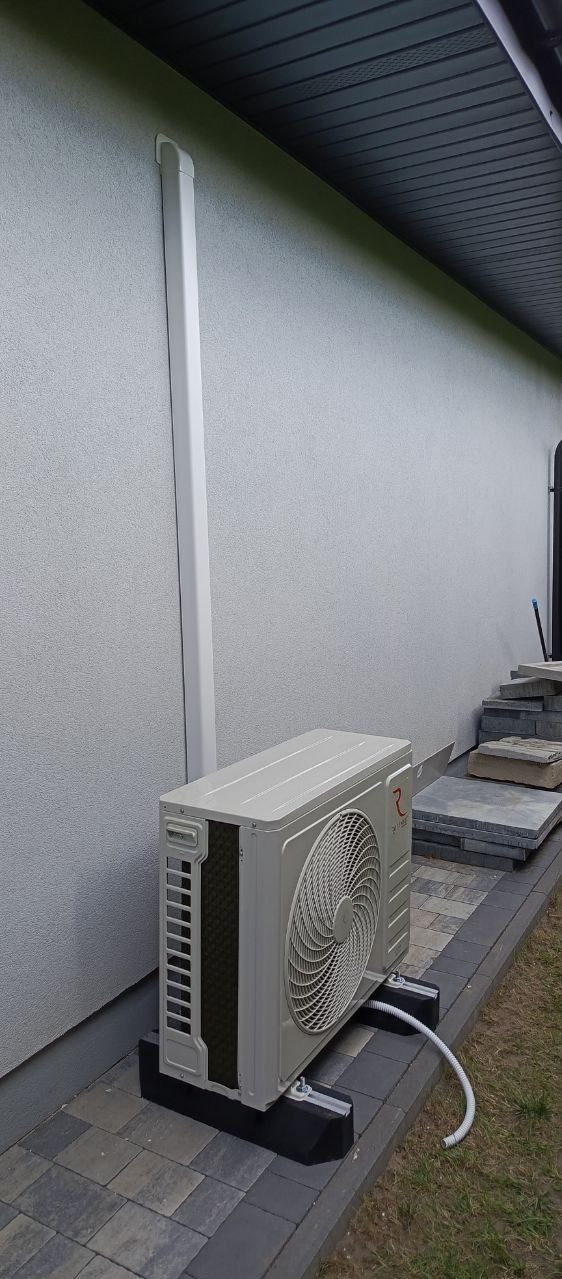 Zewnętrzna jednostka klimatyzacji zamontowana na betonowej podstawie, z widoczną rurą odprowadzającą skropliny, umieszczona na kostce brukowej przy ścianie budynku z rurą spustową.