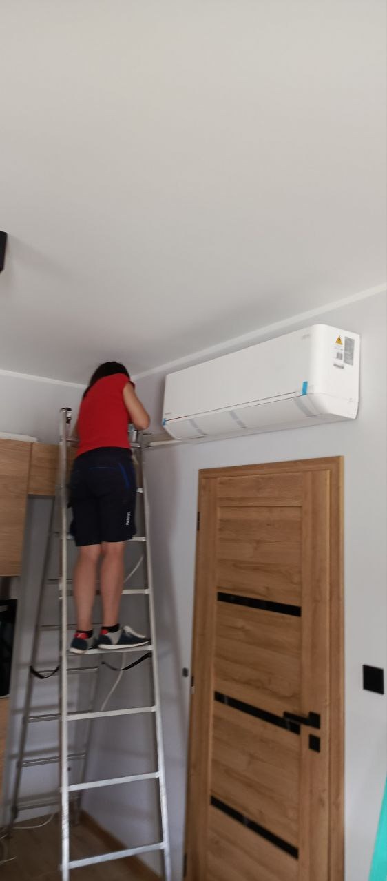 Osoba na aluminiowej drabinie instaluje biały klimatyzator ścienny w pomieszczeniu z drewnianymi elementami wystroju.