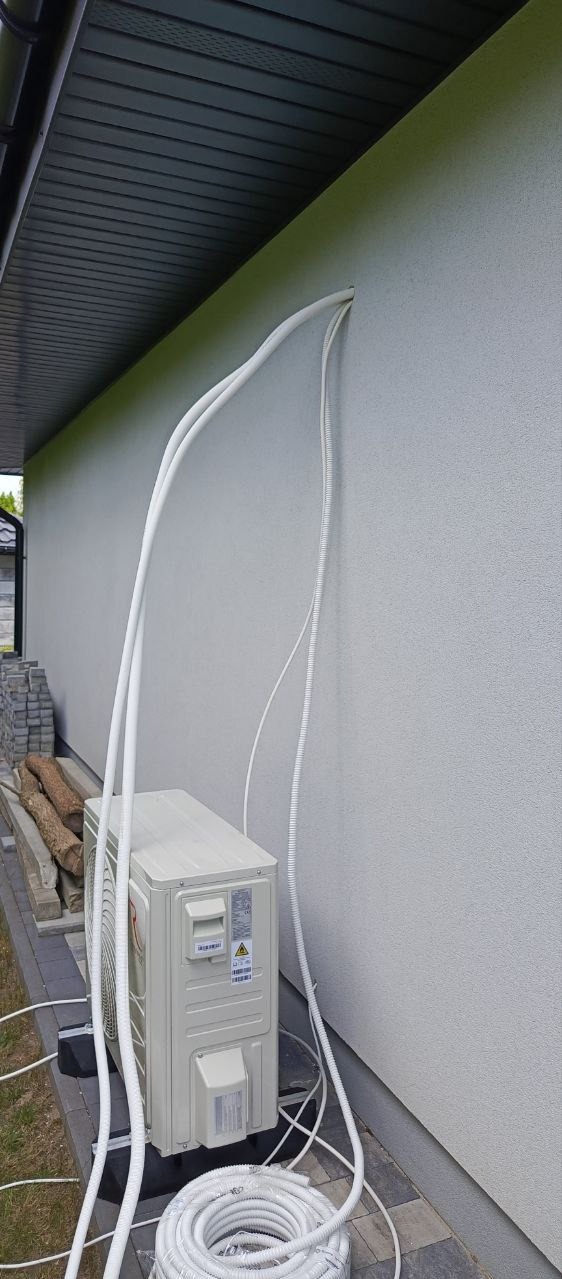 Zewnętrzna jednostka klimatyzacji na czarnych podkładkach, z podłączonymi przewodami w białych peszlach, prowadzącymi do otworu w elewacji budynku, obok ułożony zwój peszla