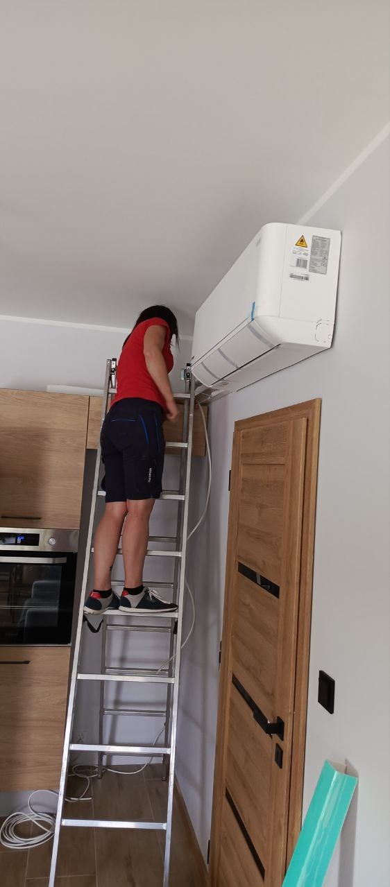 Kobieta na aluminiowej drabinie montuje biały klimatyzator ścienny w pomieszczeniu z drewnianą zabudową kuchenną i drzwiami.