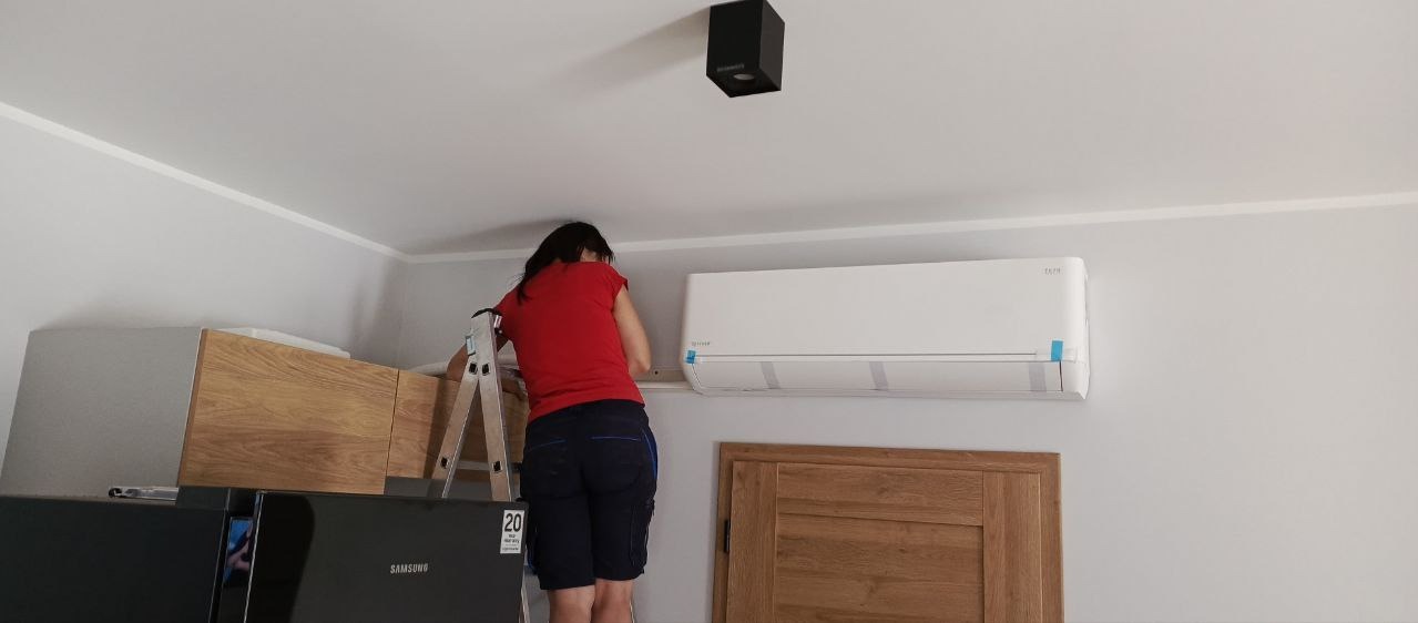Kobieta na drabinie montuje biały klimatyzator na ścianie w jasnym pomieszczeniu, widoczne zabezpieczenia na urządzeniu, czarna lampa sufitowa w tle.