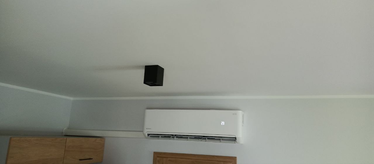 Biały klimatyzator ścienny z wyświetlaczem temperatury, zamontowany w pomieszczeniu w Tomaszowie Mazowieckim, widoczne fragmenty mebli i oświetlenie sufitowe.