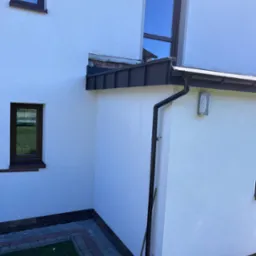 Fragment ściany budynku z nowym, ciemnym orynnowaniem i fragmentem dachu; widoczne okna z brązowymi ramami oraz kostka brukowa wokół budynku.