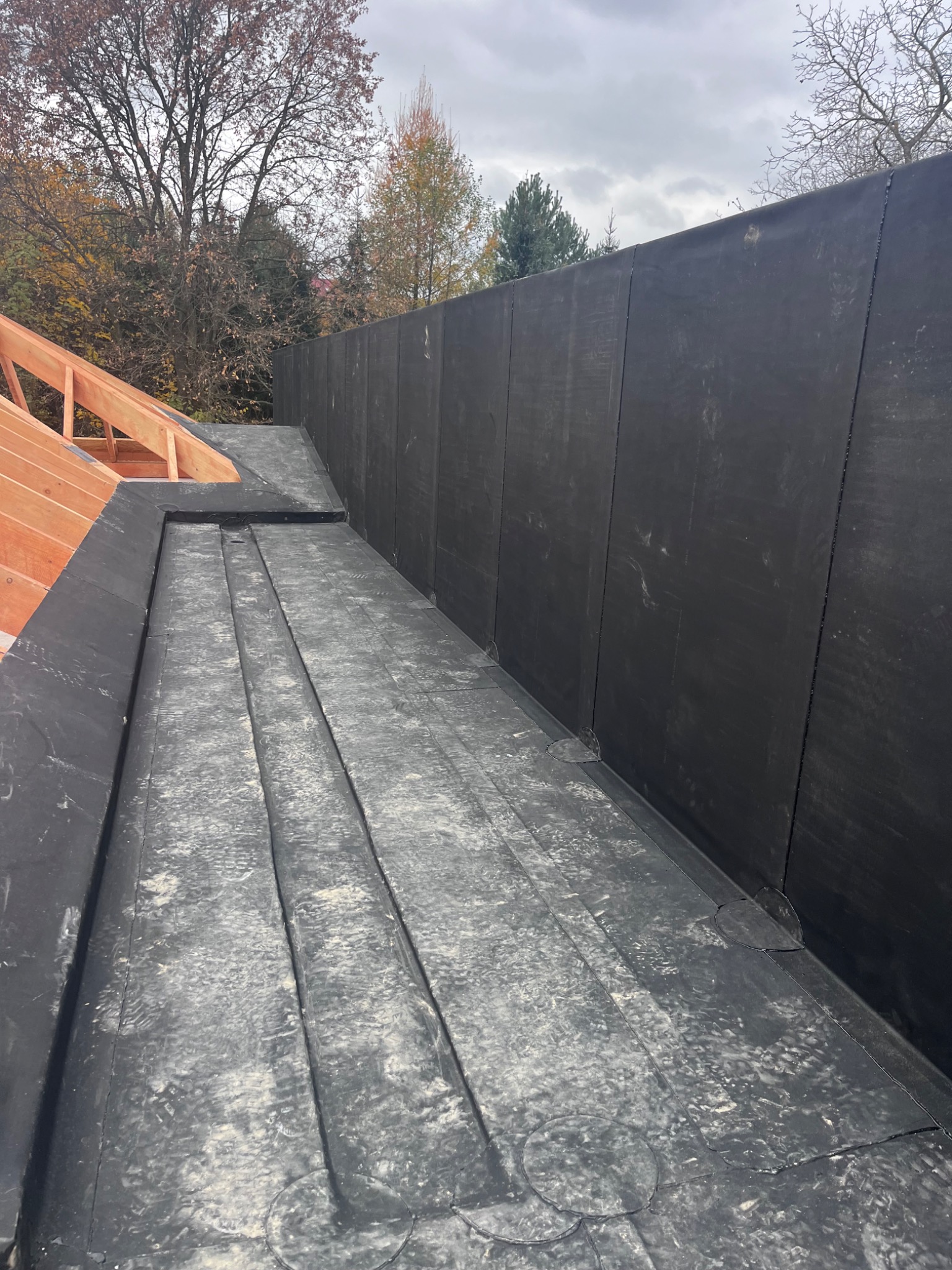Widok z góry na fragment dachu pokrytego czarną papą z widocznymi zakładkami i elementami drewnianej konstrukcji w tle, na tle drzew bez liści i pochmurnego nieba.