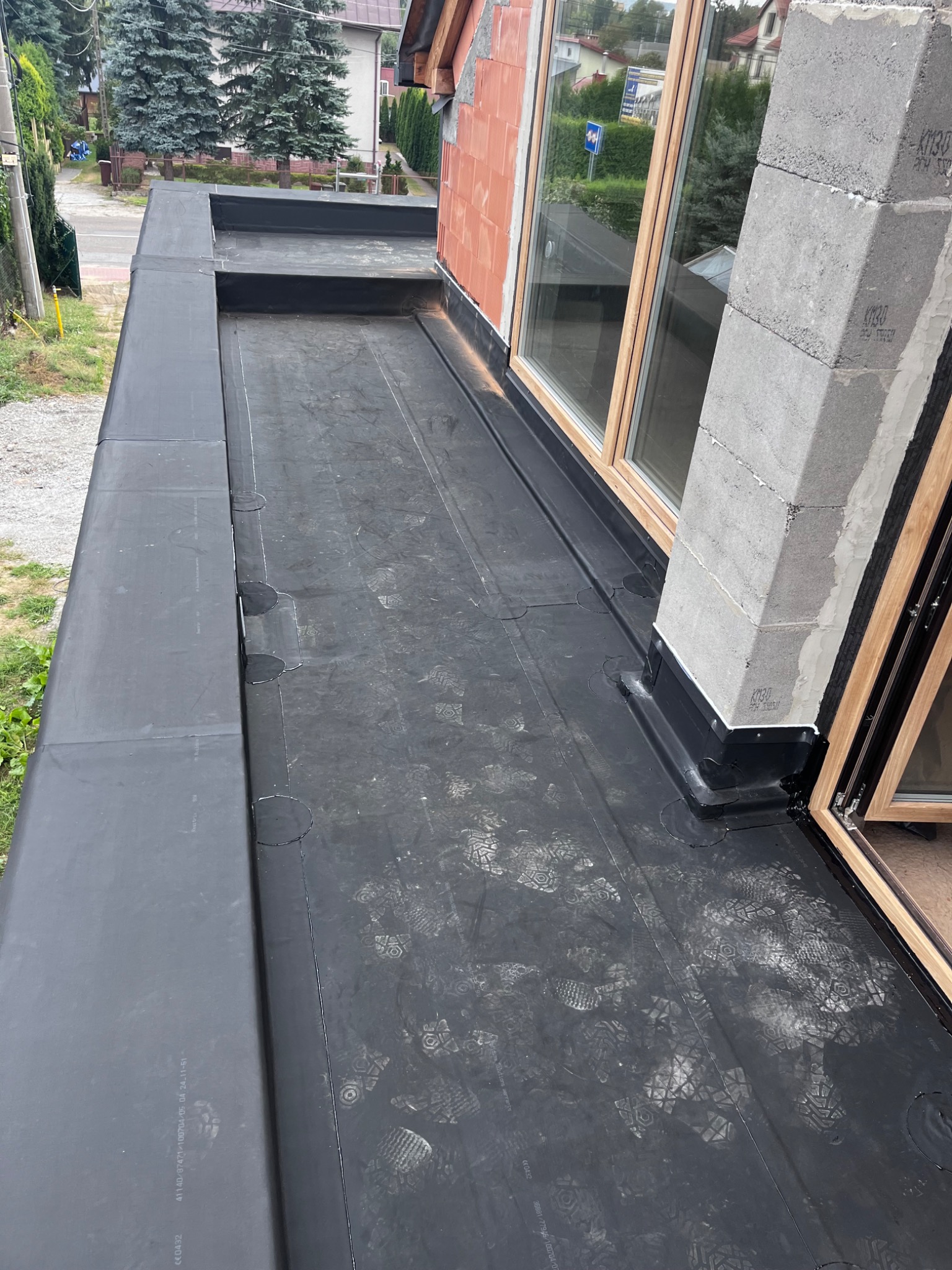 Płaski dach pokryty czarną papą, widoczne ślady butów, fragment okna i muru z pustaków betonowych.
