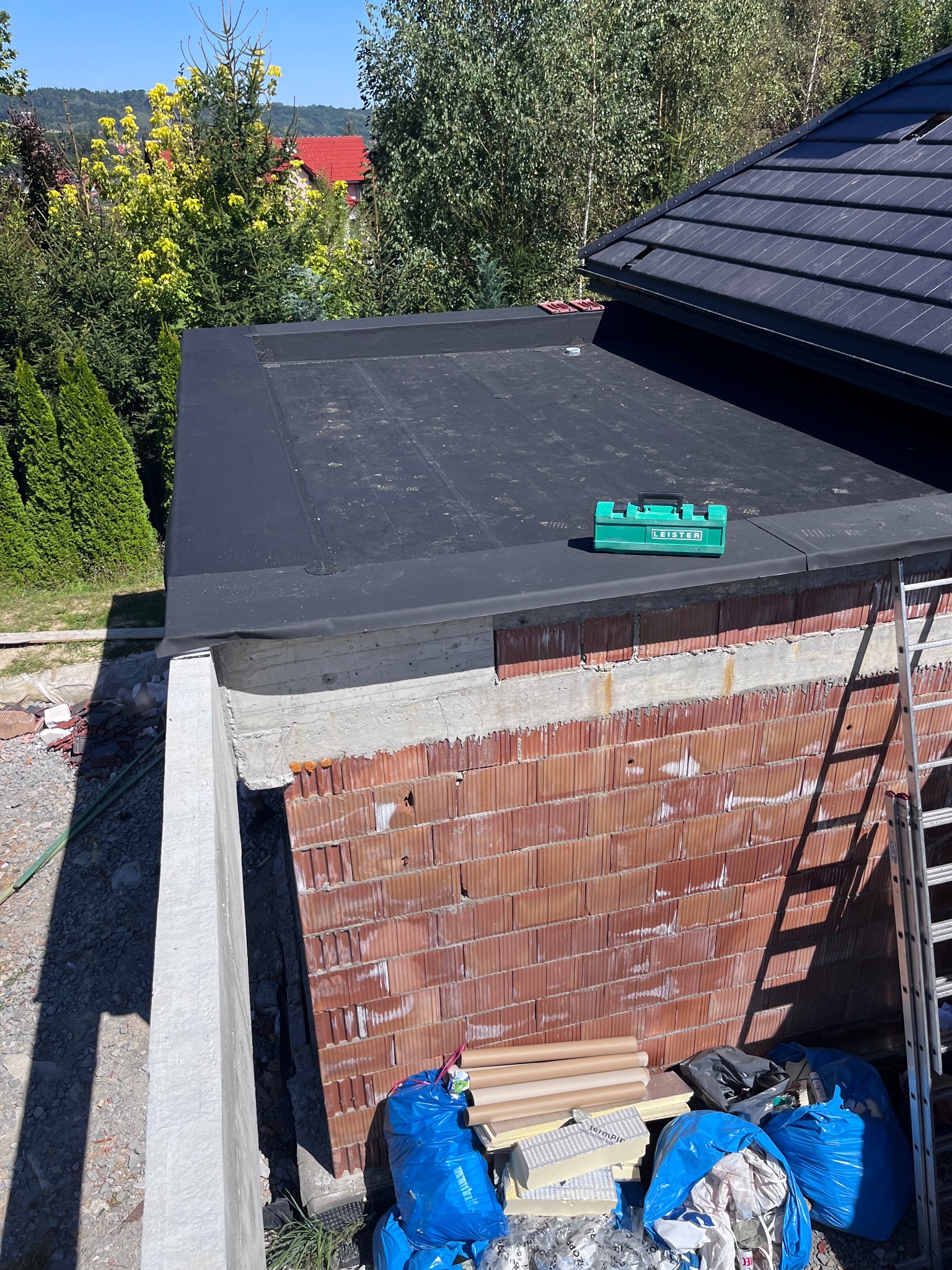 Nisko położony dach pokryty czarną papą, obok stoi zielona skrzynka narzędziowa Leister, widoczna ściana z czerwonej cegły i drabina oparta o budynek, worki ze śmieciami i materiały budowlane w tle.