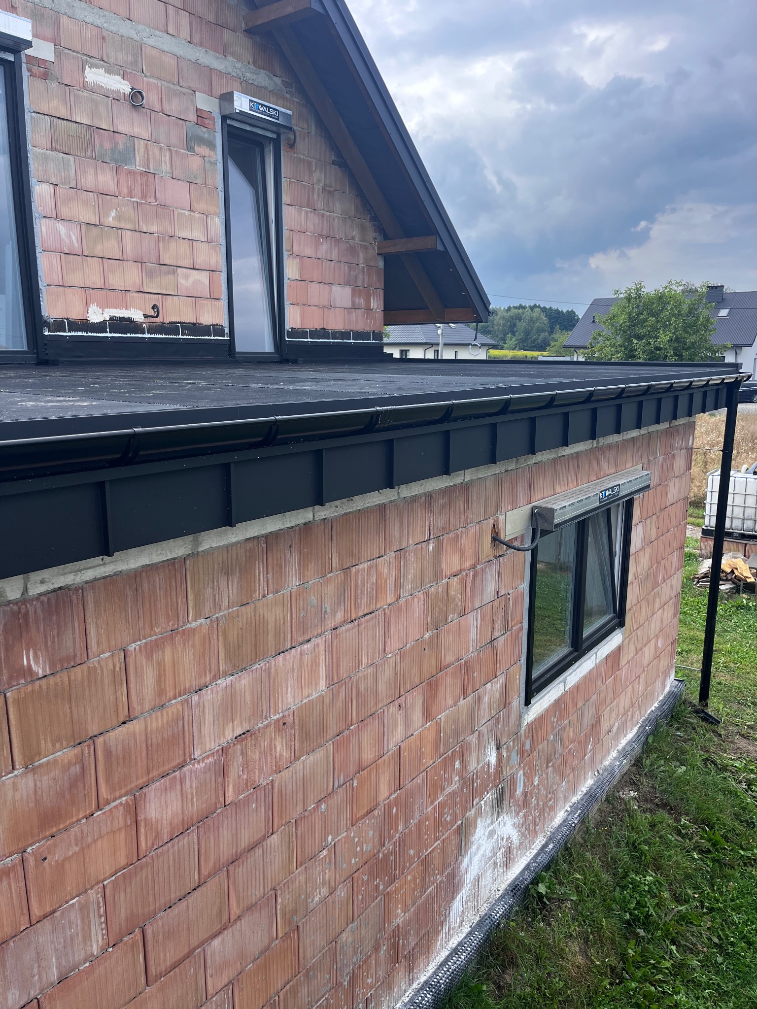 Surowy stan dachu z czarnym pokryciem i orynnowaniem na budynku z cegły ceramicznej, widoczne okna dachowe z roletami zewnętrznymi.