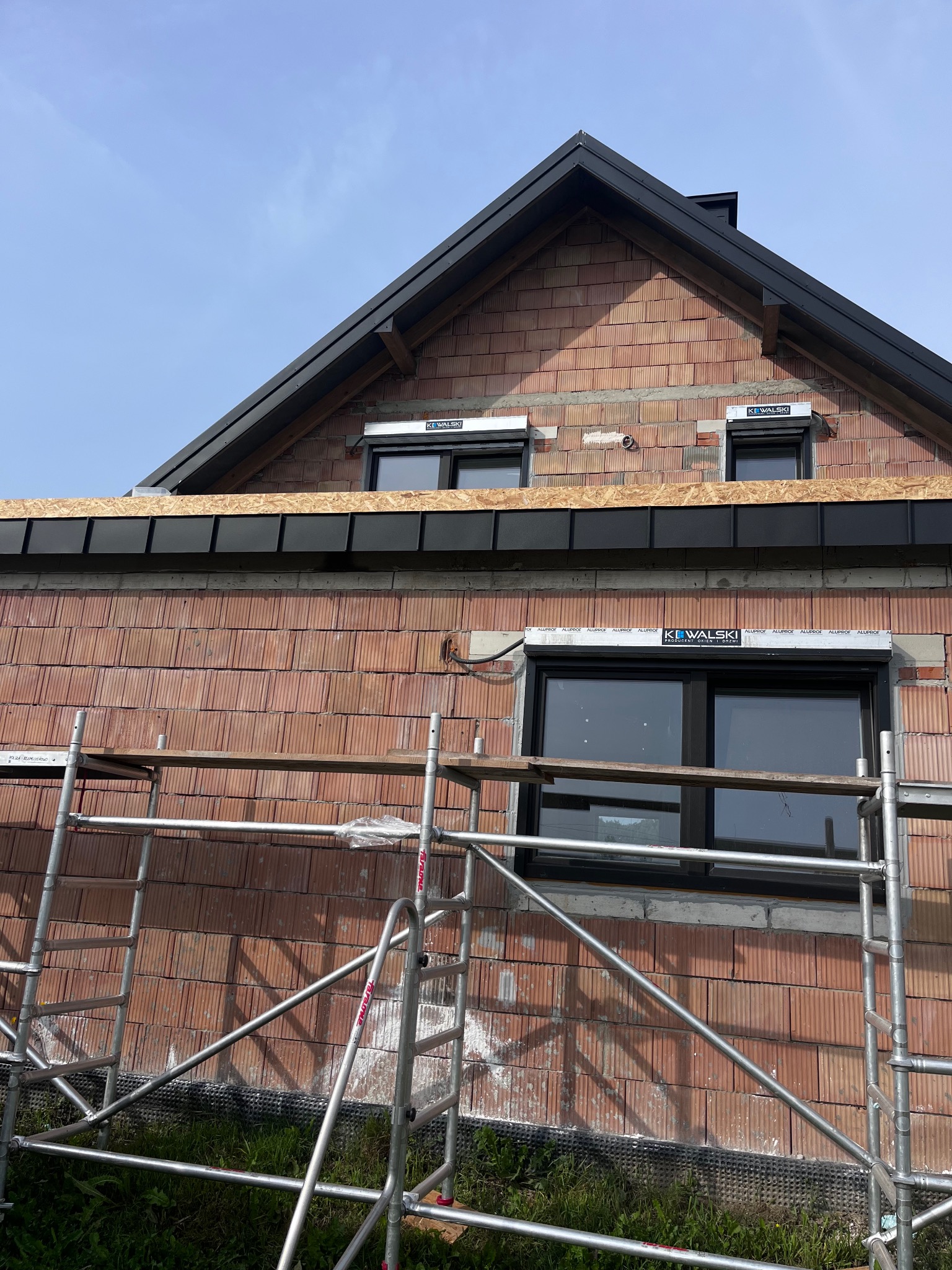 Dom w trakcie budowy z widocznym fragmentem dachu z czarną blachą, szarymi rusztowaniami i oknami z roletami zewnętrznymi.