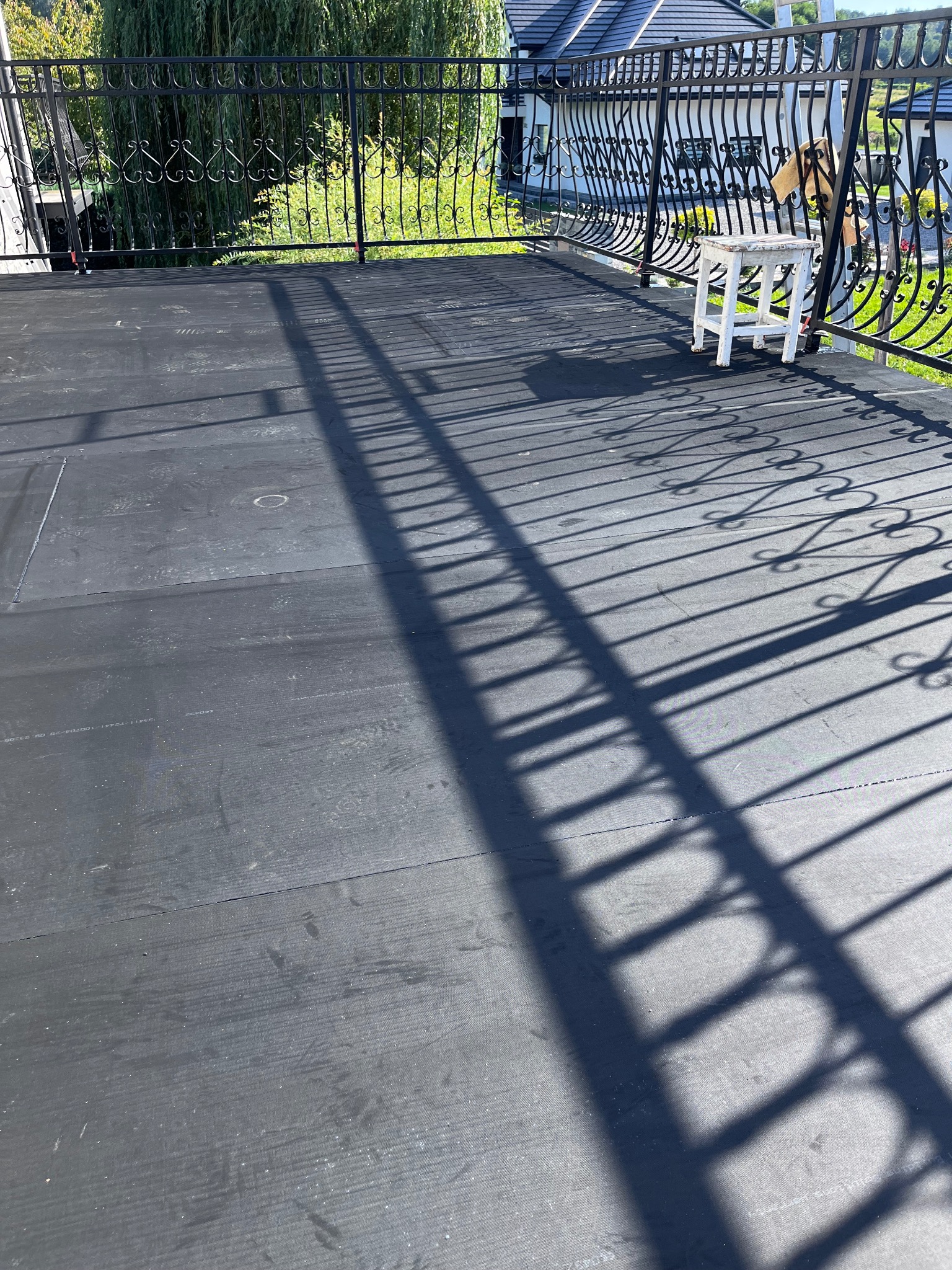 Czarna, płaska powierzchnia dachu z widocznymi łączeniami papy, z dekoracyjną, metalową balustradą i rzucanym przez nią cieniem. W tle widoczny dom i zieleń.