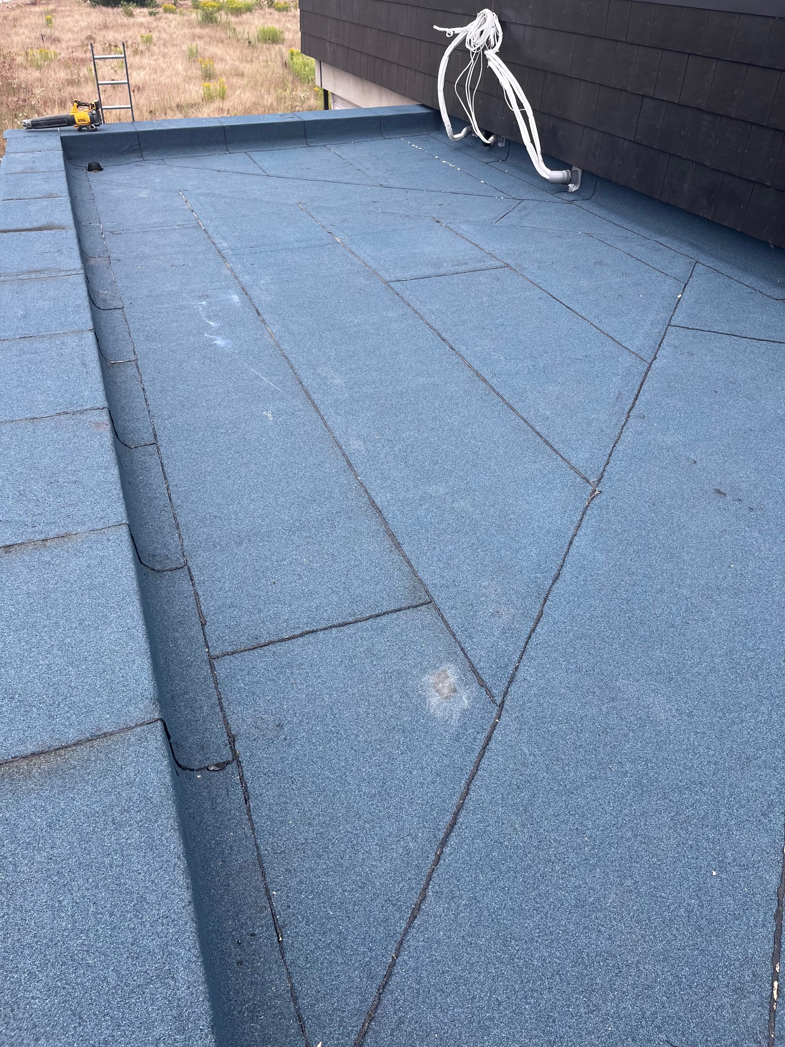 Płaski dach pokryty niebieską papą bitumiczną, widoczne łączenia płyt, elementy instalacji elektrycznej oraz drabina w tle.