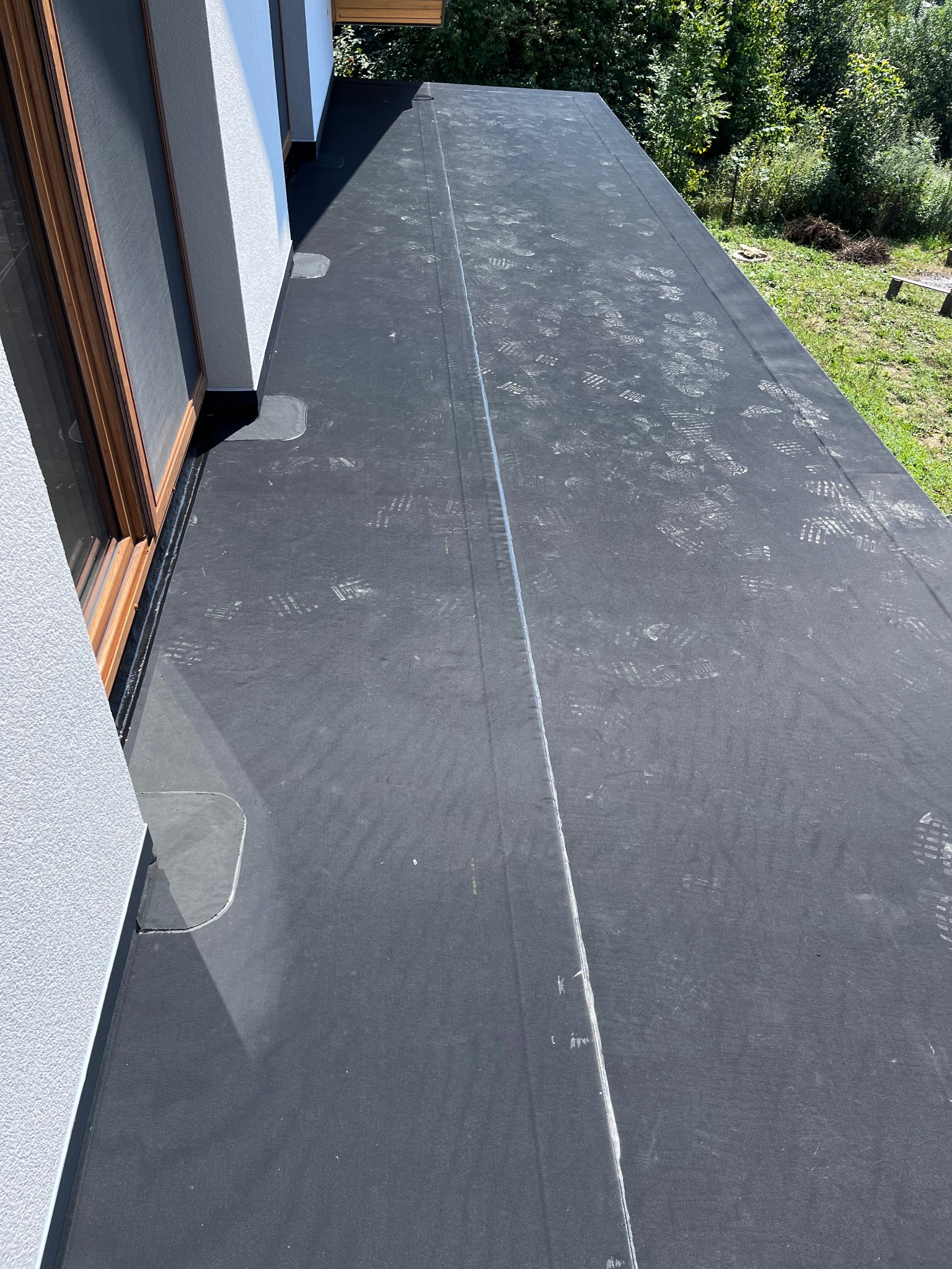 Czarna membrana dachowa na płaskim dachu z widocznymi śladami butów, złączenia zabezpieczone, obok okno dachowe i roślinność.