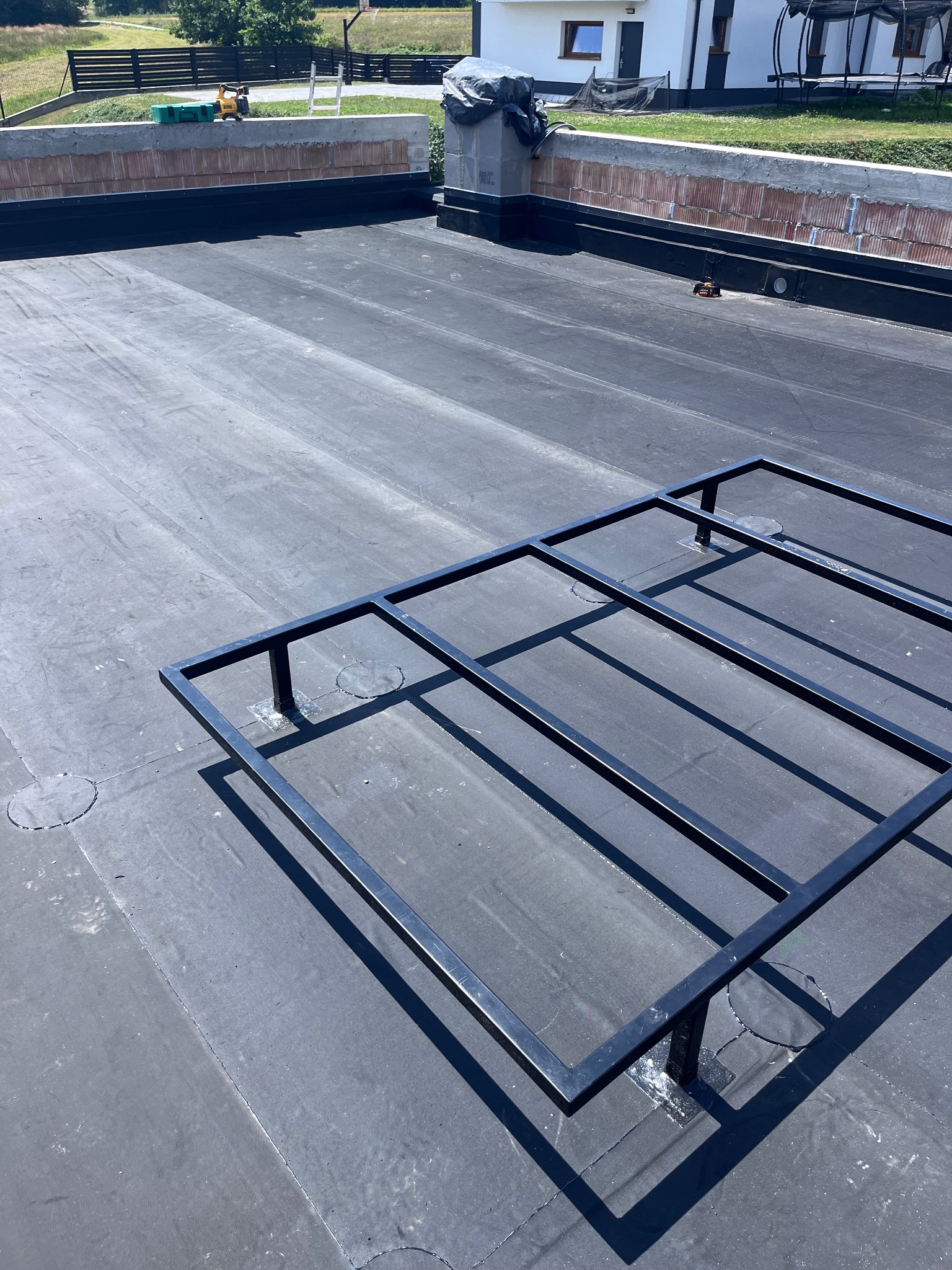 Czarna papa na płaskim dachu z zamontowaną metalową konstrukcją pod przyszłe instalacje, widoczny fragment ściany attyki z cegieł i narzędzia dekarskie w tle.