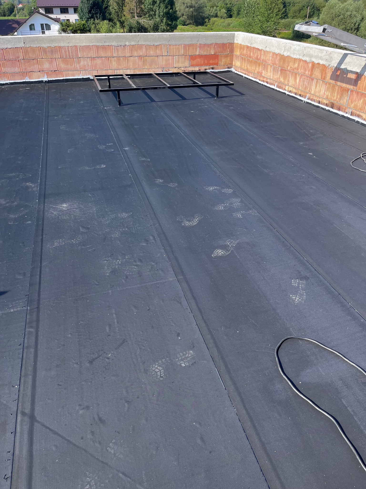 Czarna membrana dachowa z widocznymi śladami butów, metalowa konstrukcja na dachu, mur z czerwonej cegły w tle, widok z góry.