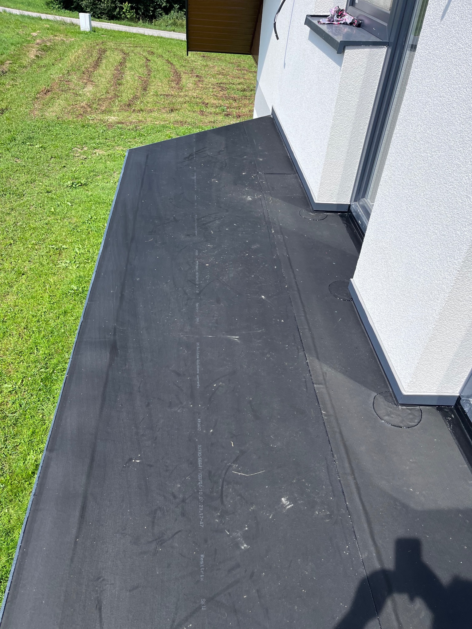 Widok z góry na płaski dach pokryty czarną membraną hydroizolacyjną, przylegający do białej ściany budynku z oknem. Widoczne detale wykończenia dachu i połączenia ze ścianą. W tle trawnik.