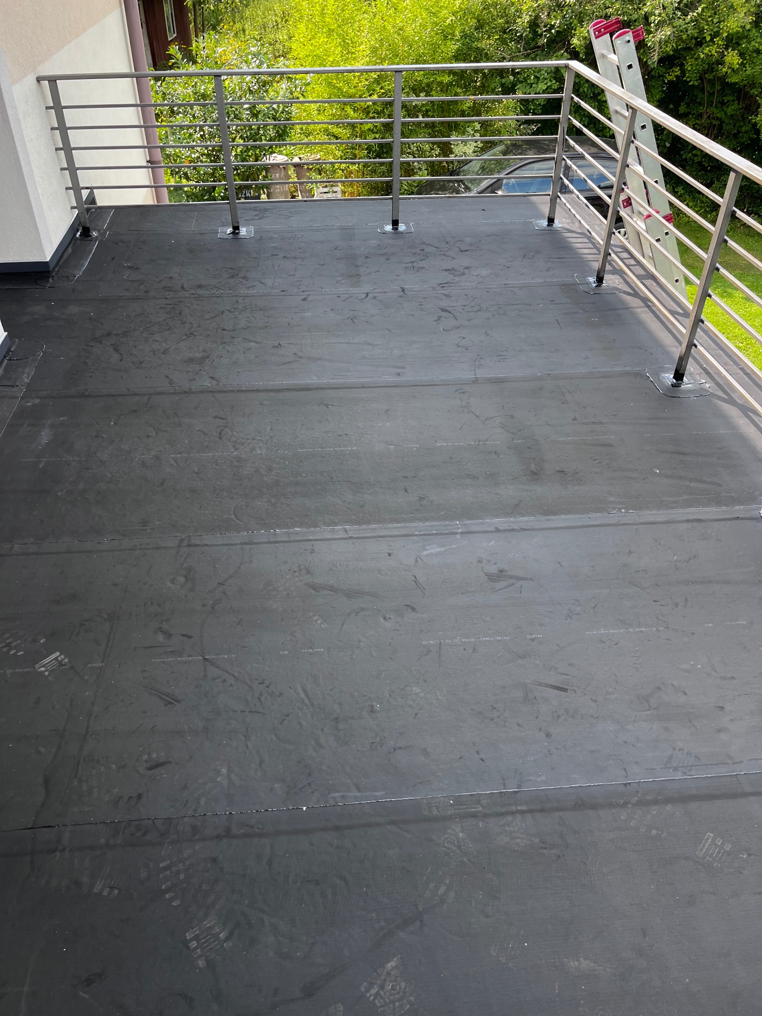 Czarna, płaska powierzchnia dachu pokryta papą z widocznymi śladami stóp, zabezpieczona metalową barierką i drabina oparta o barierkę.