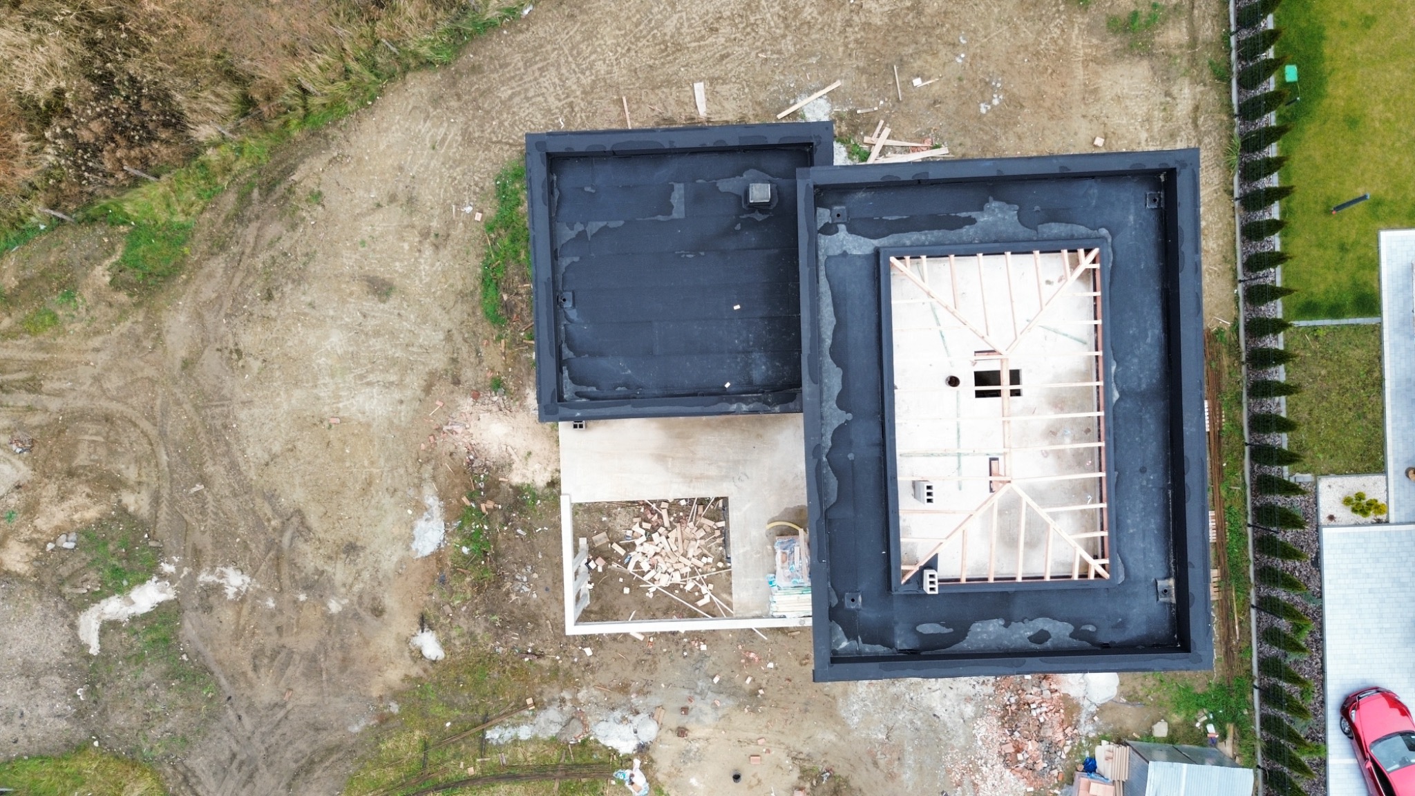 Widok z góry na płaski dach budynku w trakcie budowy, z widoczną konstrukcją drewnianą i fragmentem pokrycia bitumicznego, otoczonego ziemią i trawnikiem.