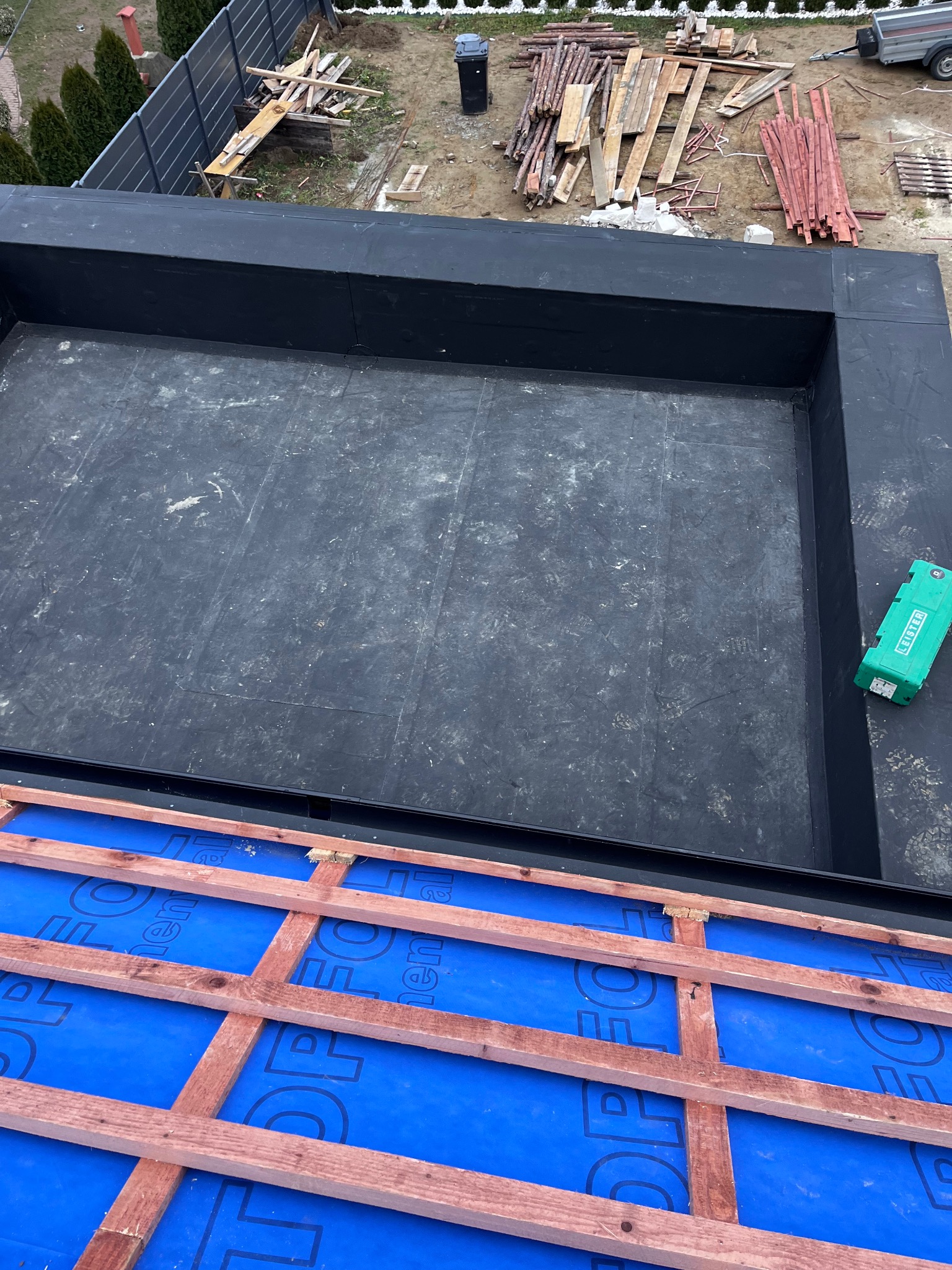 Częściowy widok dachu z hydroizolacją w kolorze niebieskim i drewnianą konstrukcją kratownicową, obok płaska powierzchnia dachu pokryta czarną membraną bitumiczną z zielonym narzędziem Leister...