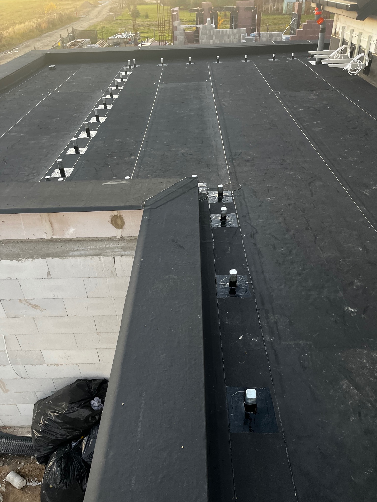 Czarna membrana dachowa na płaskim dachu budynku w trakcie budowy, widoczne mocowania i elementy konstrukcyjne. Ujęcie z góry, w tle cegły i elementy konstrukcyjne.