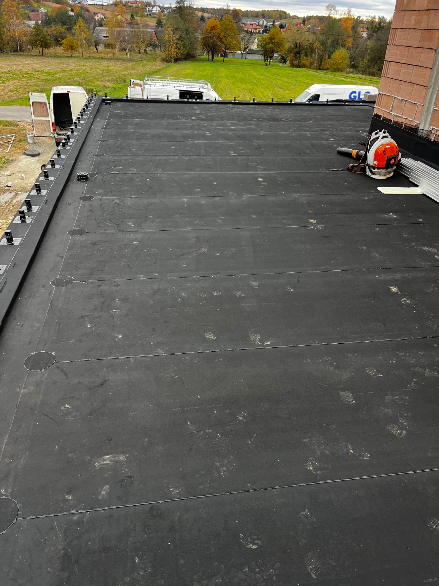 Czarna, płaska powierzchnia dachu z widocznymi łączeniami papy, wentylator i fragment ściany komina. W tle zabudowa jednorodzinna i pojazdy dostawcze.