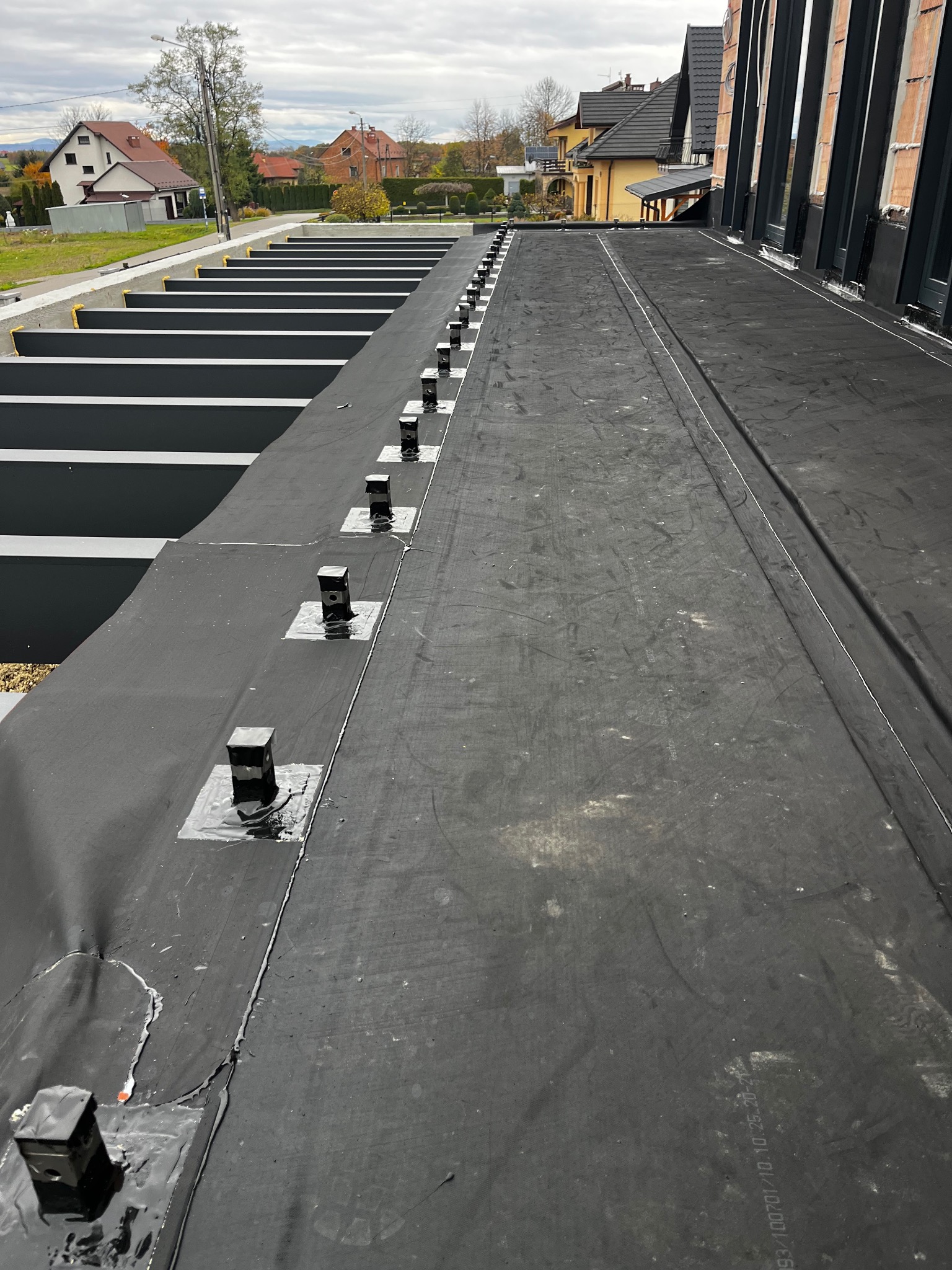 Czarna membrana dachowa z zamocowanymi elementami, widok z góry na płaski dach z elementami konstrukcyjnymi i zabudową w tle. Detal hydroizolacji.