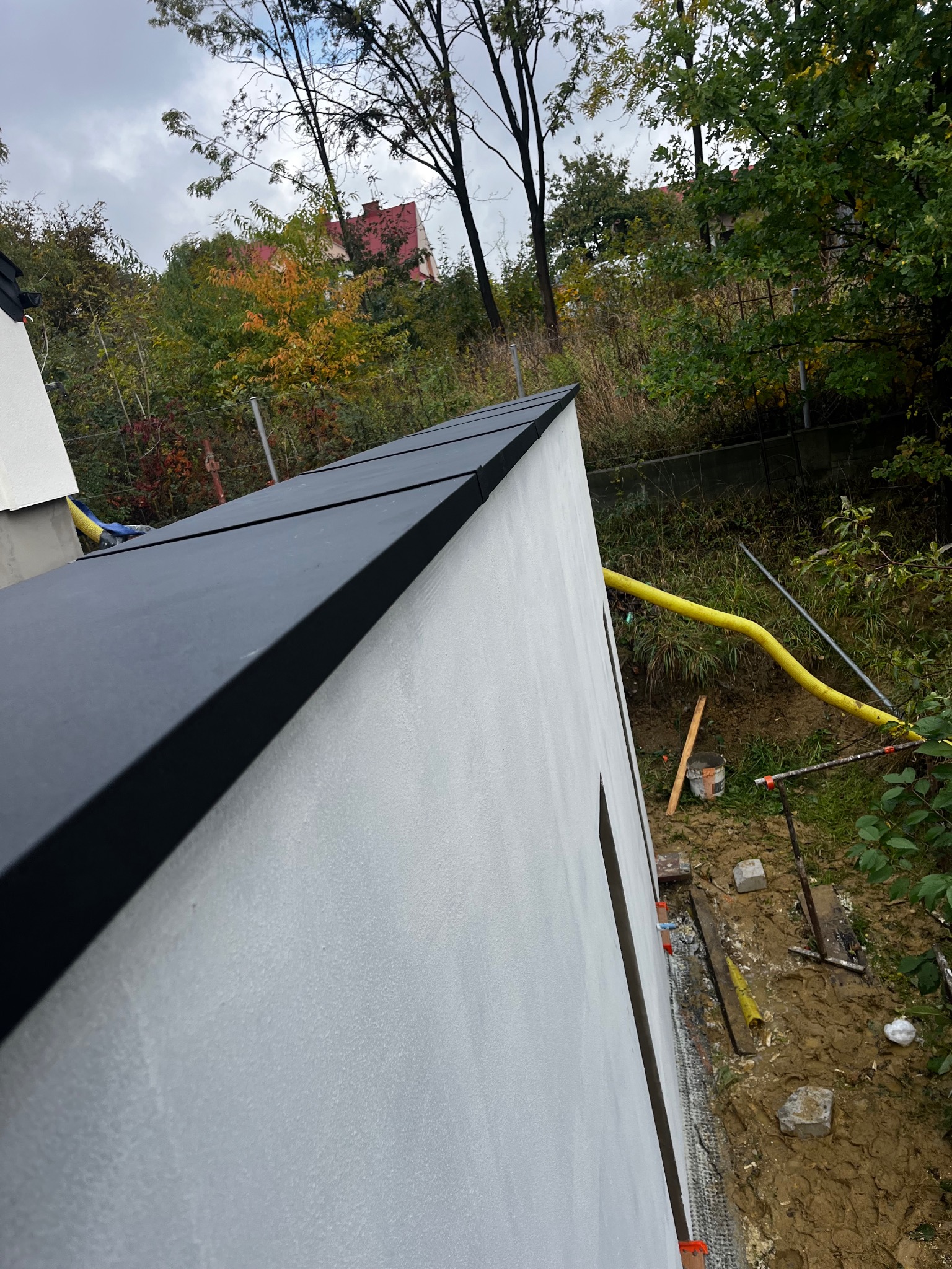 Nowoczesny, płaski dach z czarną blachą na wykończonym budynku. Widoczny fragment elewacji, rynna i otoczenie z zielenią. Ujęcie z dołu, podkreślające geometrię dachu.