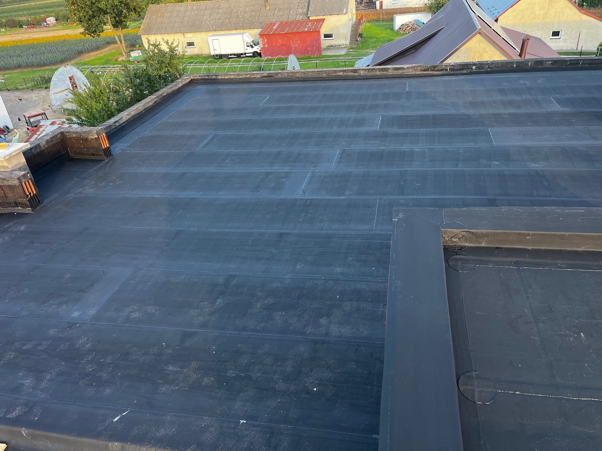 Czarny, płaski dach pokryty papą termozgrzewalną z widocznymi śladami butów, na tle zabudowań gospodarczych i pola uprawnego w słoneczny dzień.