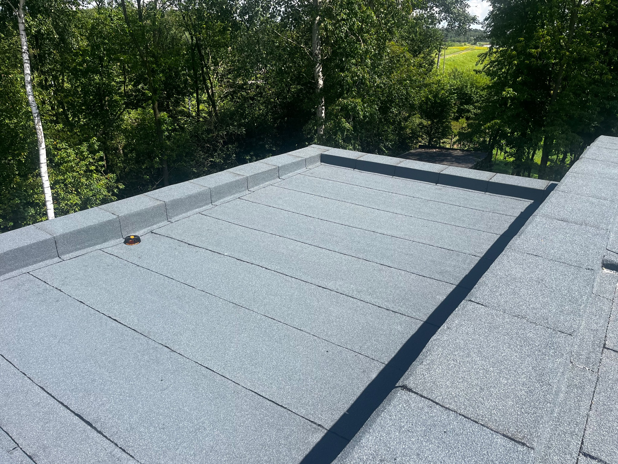 Szary dach pokryty papą termozgrzewalną na tle zieleni drzew. Widoczny fragment obróbki blacharskiej i kominek wentylacyjny. Słoneczny dzień.