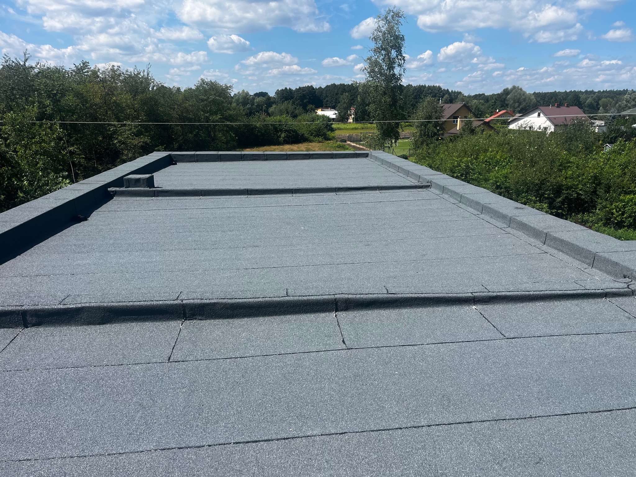 Szary, płaski dach pokryty papą termozgrzewalną, z widocznymi obróbkami blacharskimi i elementami wentylacyjnymi na tle zieleni i błękitnego nieba.
