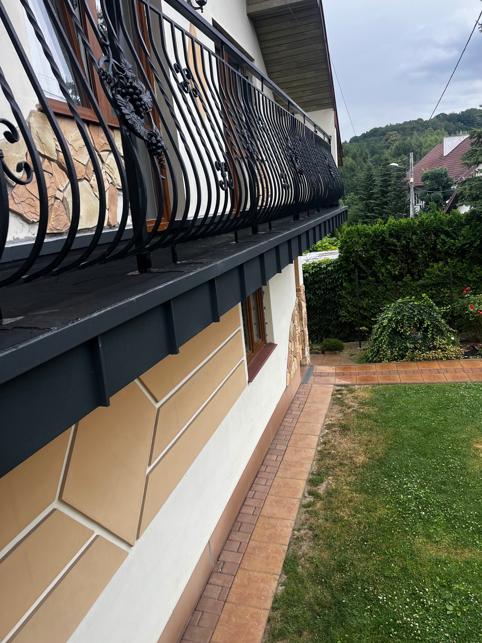 Elegancki balkon z kutą balustradą zdobioną motywem winogron, przylegający do budynku z dekoracyjną elewacją w odcieniach beżu i brązu. Widok z boku, na tle zieleni.