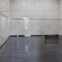 Wykończona ściana kabiny prysznicowej z białymi płytkami z motywem kwiatowym i szarymi płytkami imitującymi kamień, z widocznym odpływem liniowym w podłodze.