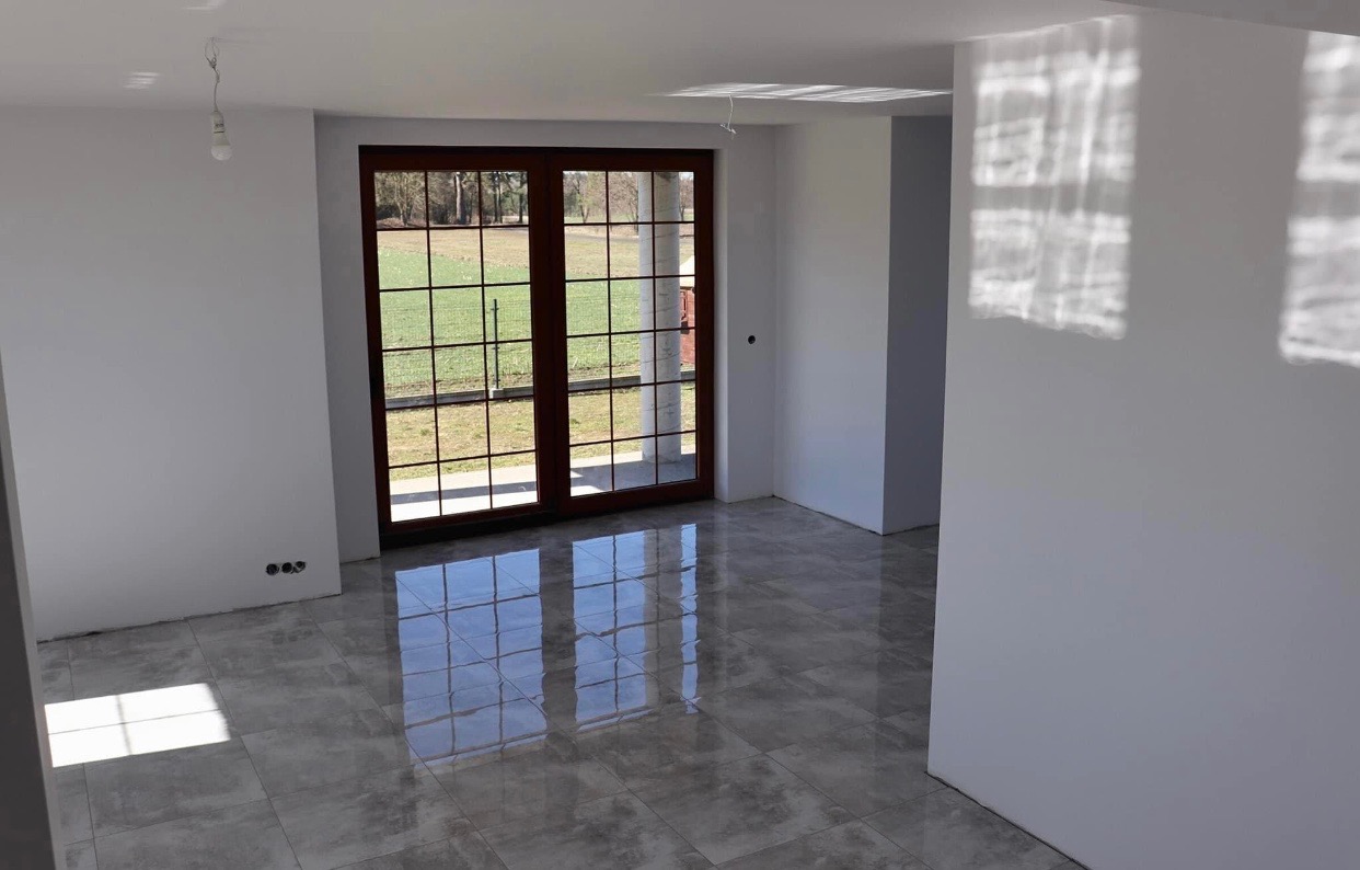 Wnętrze po remoncie: szare płytki podłogowe z połyskiem, białe ściany z widocznymi refleksami światła, brązowe drzwi balkonowe z widokiem na zewnątrz, surowa żarówka zwisająca z sufitu.